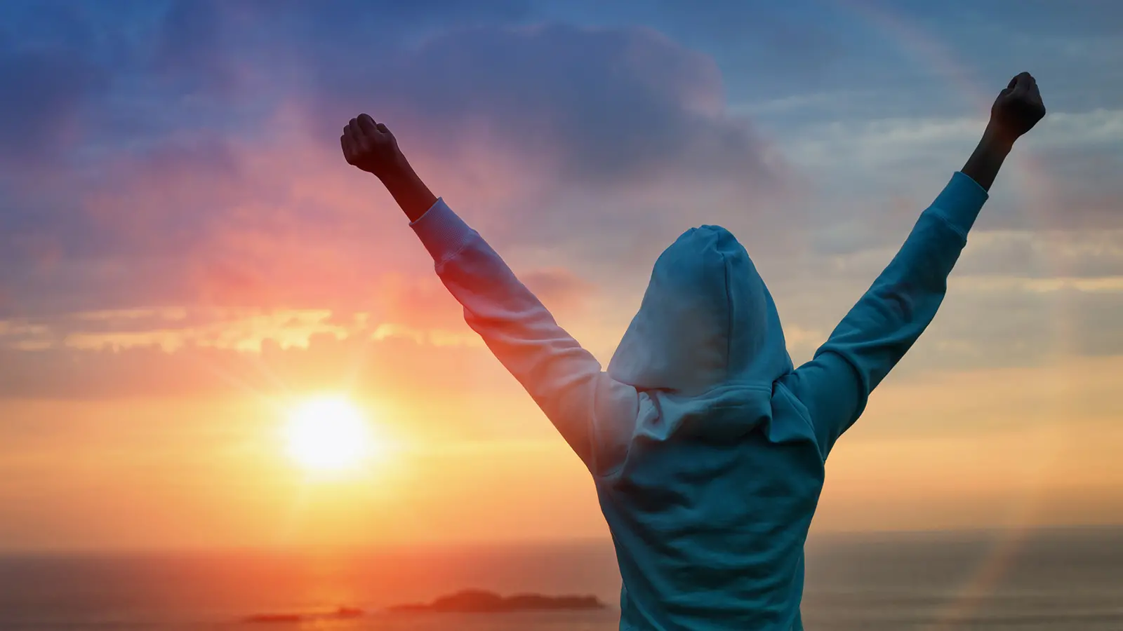 Person in hooded sweatshirt with arms raised in victory staring at sunrise