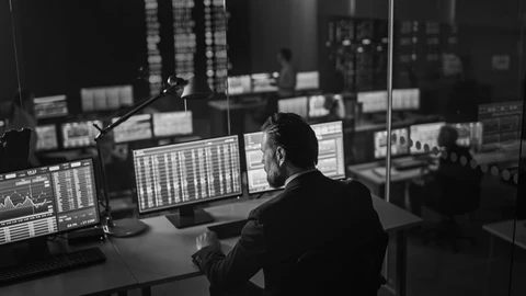 Grayscale photo of man sitting in room with multiple monitors displaying charts and data
