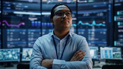 Gentlmen in button up shirt with arms crossed looking confident  with monitors in background.