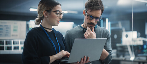 1 person holding a laptop while another one stands beside while reviewing something being presented on laptop. 